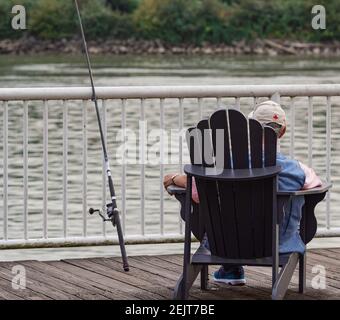 Vista posteriore di un vecchio uomo che pesca sul molo. Un anziano si siede su una sedia di legno vicino al fiume Fraser a New Westminster. Stile di vita attivo. Via Foto Stock