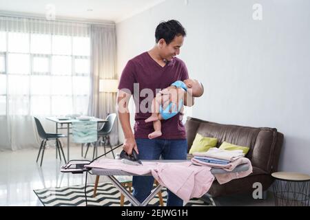 ritratto di padre asiatico che stira i suoi vestiti mentre lo tiene bambino neonato sulla sua mano Foto Stock