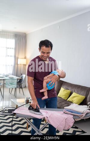 ritratto di padre asiatico che stira i suoi vestiti mentre lo tiene bambino neonato sulla sua mano Foto Stock