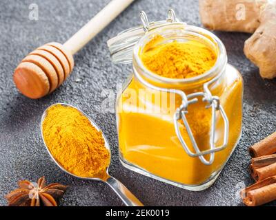 Polvere curcuma essiccata in vasetto di vetro e cucchiaio su fondo cementizio nero. Ingredienti per latte dorato, latte curcuma o bevanda detox - cucchiaia di miele, radice di zenzero, bastoncini di cannella e anice stellato Foto Stock