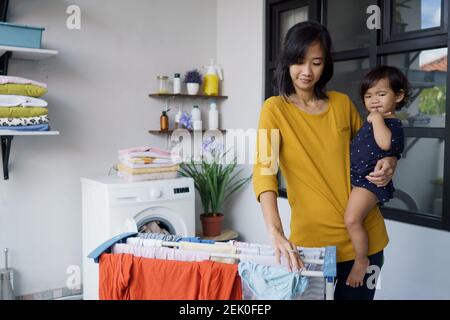 madre asiatica una casalinga che asciuga e appende i vestiti in loundry stanza a casa mentre porta il suo bambino Foto Stock