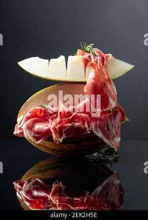 Prosciutto con melone e rosmarino su fondo nero riflettente. Foto Stock