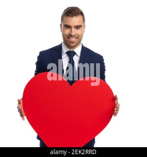 elegante uomo bearded in tuta blu navy che tiene grande cuore rosso per il giorno di san valentino e sorridente isolato su sfondo bianco in studio, ritratto Foto Stock