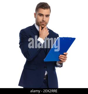 giovane uomo pensivo su sfondo bianco con vestito blu navy, tenendo la mano al mento e pensando, in piedi isolati in studio Foto Stock