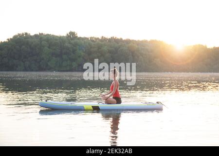 Donna che medita e pratica yoga durante l'alba su una tavola da paddleboard Foto Stock