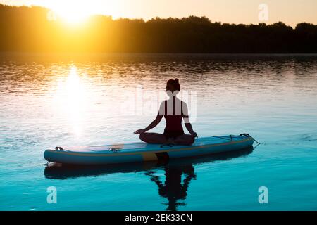 Donna che medita e pratica yoga durante l'alba su una tavola da paddleboard Foto Stock