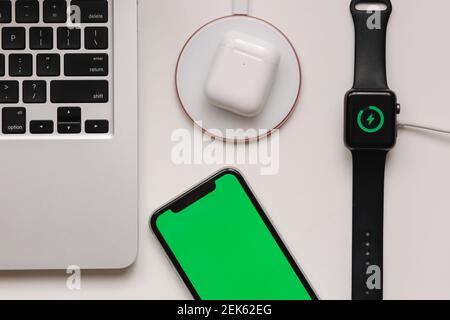 Luogo di lavoro con computer portatile su tavolo e dispositivi. Ricarica wireless di Smart Watch e cuffie wireless su tavolo bianco. Smartphone con schermo verde Foto Stock