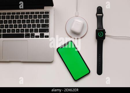 Luogo di lavoro con computer portatile su tavolo e dispositivi. Ricarica wireless di Smart Watch e cuffie wireless su tavolo bianco. Smartphone con schermo verde Foto Stock