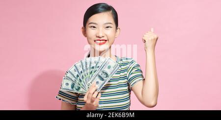 Felice giovane donna facendo gesto vincitore, tenendo fan di denaro in contanti in banconote in dollari isolato su sfondo rosa Foto Stock