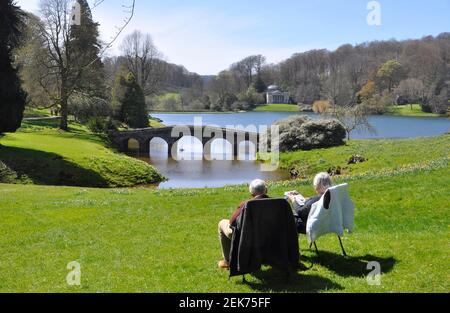 Una coppia che si siede godendo il sole primaverile mentre guarda attraverso il lago e il suo Ponte Palladiano verso il Pantheon presso i Giardini Stourhead, Wiltsh Foto Stock