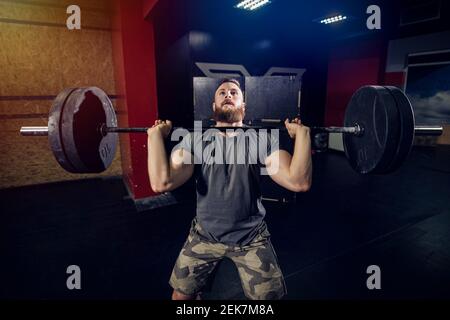 Uomo robusto e sportivo che solleva il peso pesante in camera oscura. Foto Stock