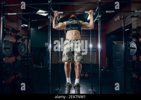 Muscoloso uomo caucasico bearded facendo pull-up in stile militare gilet ponderato in palestra crossfit. Piastre di peso, kettlebells, barbell e pneumatici crossfit Foto Stock