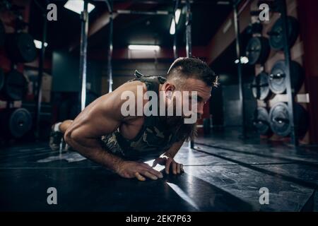 Uomo caucasico muscoloso bearded facendo push-up in stile militare gilet ponderato in palestra. Piastre per pesi sullo sfondo. Foto Stock