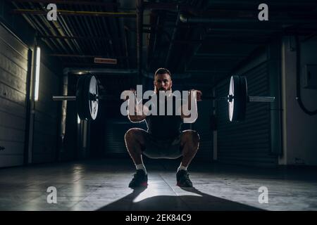 Muscoloso attraente uomo caucasico bearded facendo squat di barbell anteriore nel corridoio sotterraneo. Unità di stoccaggio e tubi sullo sfondo. Foto Stock