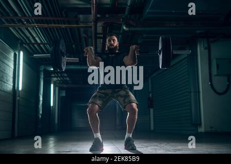 Uomo forte bearded che fa l'allenamento, in piedi e sollevando barbell all'interno. Unità di stoccaggio e tubi sullo sfondo. Foto Stock