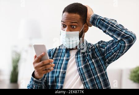 Guy scioccato che usa il telefono indossando la maschera di faccia seduto sul posto di lavoro Foto Stock