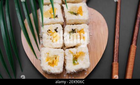 Sushi da dessert. Kiwi dolce, panini di sushi di ananas. Sushi su un vassoio di legno su sfondo nero con una foglia tropicale Foto Stock