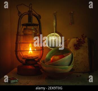 Una lampada a cherosene che brucia illumina una mela in una ciotola di ceramica, un colander e una chiave sulla parete, un libro con un ramo di rowan. Vintage. Retrò. Foto Stock