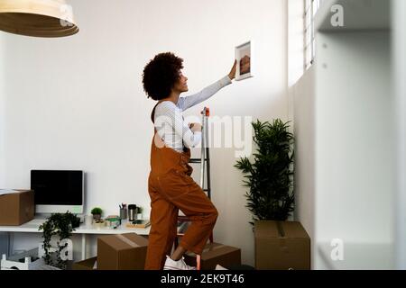 Donna afro che mette la cornice del quadro sulla parete mentre si arrampica sopra scala in appartamento nuovo Foto Stock