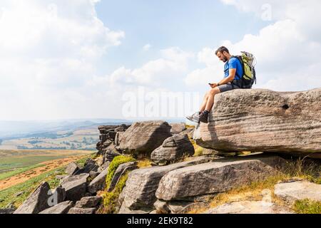 Giovane escursionista maschile che utilizza lo smartphone mentre si siede in cima di montagna durante le vacanze Foto Stock