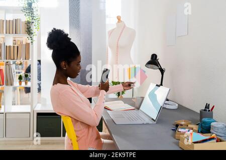 Donna d'affari che scatta foto di Swatch di tessuto attraverso il telefono cellulare mentre seduto in ufficio a casa Foto Stock