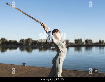 Giovane donna che fa arti marziali con la spada nel parco vicino lago in giornata di sole Foto Stock
