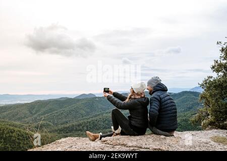 Coppia che prende selfie mentre si siede sul punto di osservazione contro il cielo Foto Stock