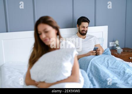 Bella ragazza giovane seduta sul letto e che guarda triste mentre i suoi fidanzati non prestano attenzione a lei. Sta sorridendo e guardando il suo telefono. Foto Stock