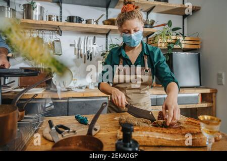 Giovane chef che indossa una maschera protettiva per il viso tagliando il salame mentre si è in piedi in cucina Foto Stock