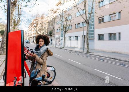 Giovane donna che guarda via mentre si sta in piedi da una macchina per biglietti a. parcheggio per biciclette in città Foto Stock