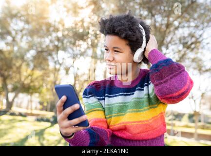 Ragazza che indossa le cuffie utilizzando il telefono cellulare mentre si è seduti con la mano nei capelli al parco Foto Stock