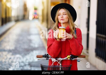 Donna bionda che indossa un cappello che beve succo mentre si trova in piedi con la bicicletta sul sentiero Foto Stock