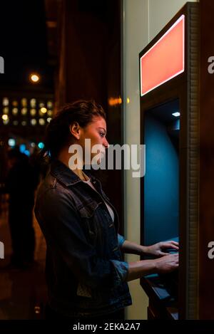Giovane donna con giacca in denim che ritira denaro da ATM in città di notte Foto Stock