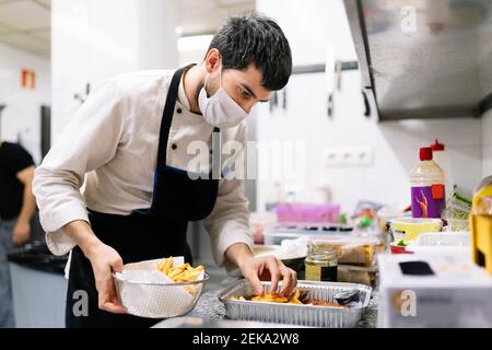 Chef maschile in maschera protettiva che dispone patatine fritte in contenitore in alluminio nella cucina del ristorante Foto Stock