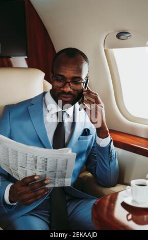 Giovane uomo d'affari che legge il giornale mentre parla con lo smartphone dentro aereo Foto Stock