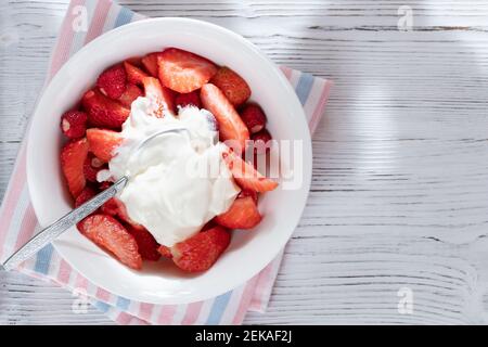 Fragole mature a fette naturali in una ciotola bianca con panna acida su fondo di legno bianco. Vista dall'alto, spazio di copia. Foto Stock