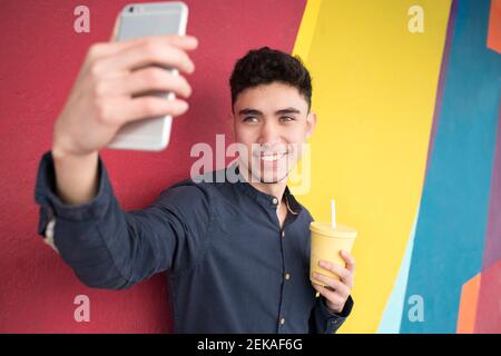 Giovane uomo che prende selfie attraverso lo smartphone mentre beve succo contro la parete multicolore Foto Stock
