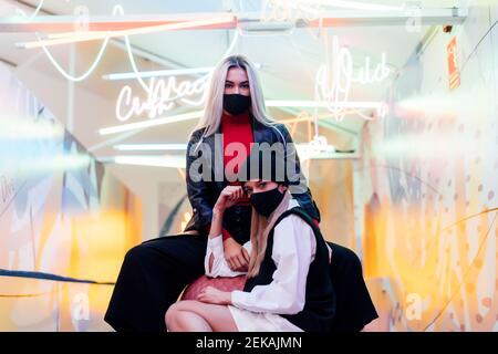 Le amiche indossano maschere nere che si pongono mentre si siedono contro parete in vetro illuminata Foto Stock
