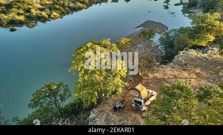 Vista aerea di una coppia accampata con la loro jeep e tenda sul tetto e facendo colazione di fronte al fiume, zona del fiume Cunene, Angola Foto Stock