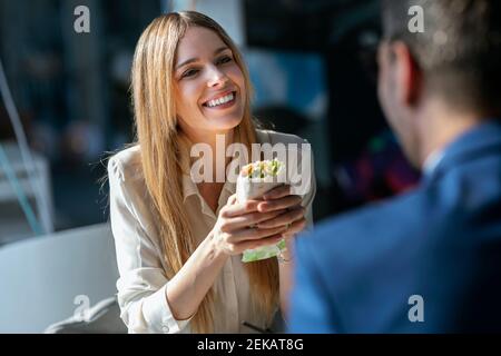 Donna d'affari che tiene il cibo mentre guarda un collega seduto in caffetteria Foto Stock