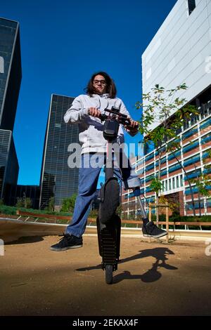 Giovane uomo disabile che pratica acrobazie su scooter elettrico a spinta dentro parco giochi contro gli edifici della città Foto Stock