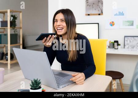 Felice studentessa con il computer portatile che parla con il telefono vivavoce mentre seduto sulla sedia a casa Foto Stock