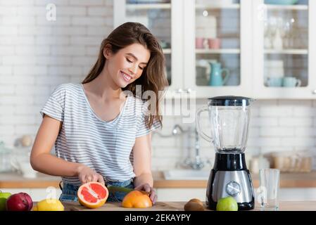 donna felice che taglia la frutta fresca vicino al frullatore in cucina Foto Stock