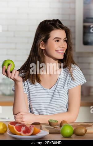 donna soddisfatta che tiene la mela matura vicino alla frutta fresca in cucina Foto Stock