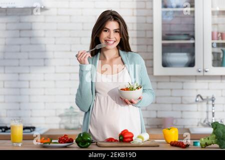 donna allegra incinta mangiare insalata vicino verdure fresche in cucina Foto Stock