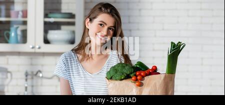 gioiosa donna che guarda la macchina fotografica vicino verdure fresche in borsa di carta, striscione Foto Stock