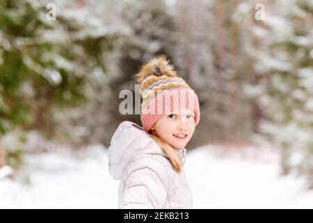 Ragazza sorridente che indossa abiti caldi in piedi in foresta durante l'inverno Foto Stock