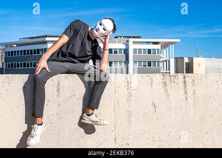 Uomo che indossa una maschera bianca che guarda lontano mentre si siede con la testa in mani sulla parete Foto Stock