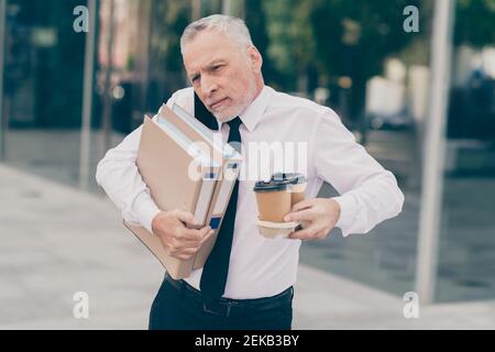 Foto di gravi capelli grigi vecchio uomo d'affari tenere carte telefono coffee talk indossare una camicia bianca all'aperto vicino al centro di lavoro Foto Stock