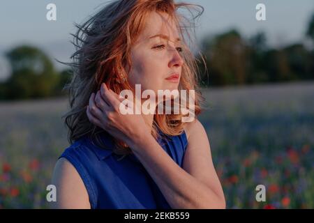 Donna premurosa con capelli aggrovigliati contemplando in campo papavero durante tramonto Foto Stock